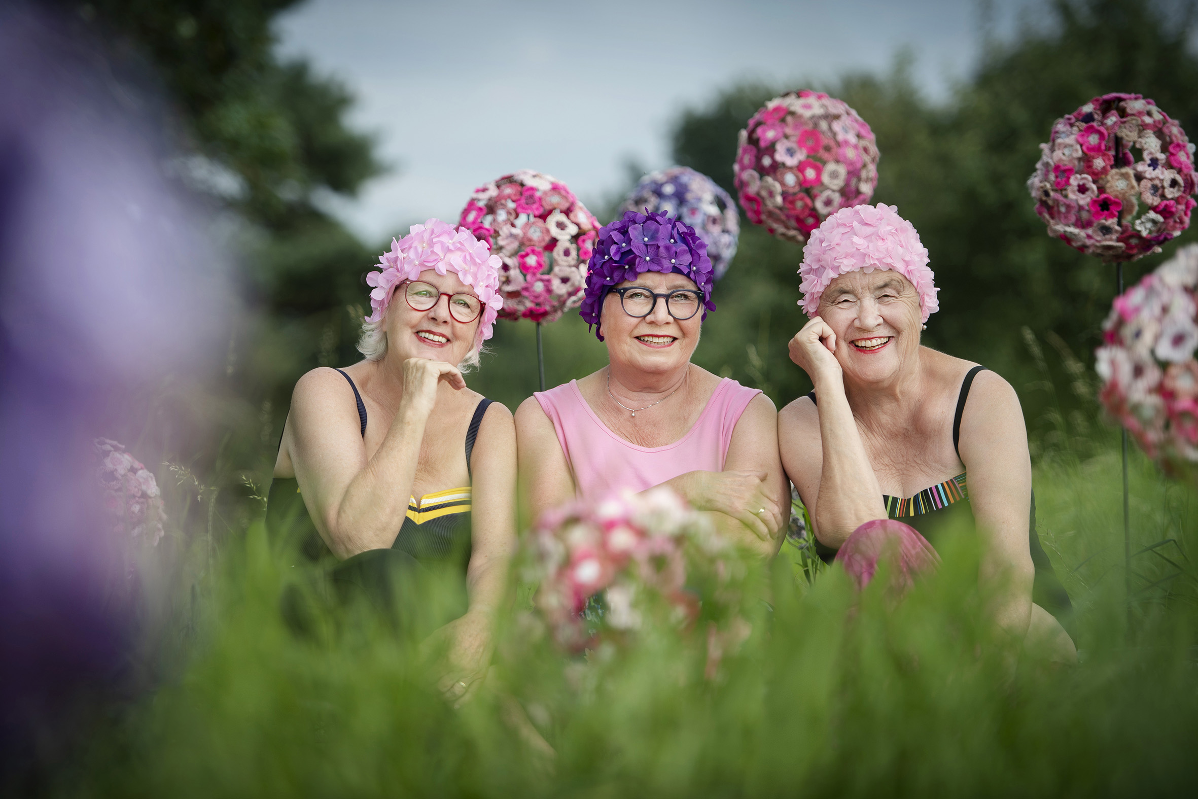 Drei Damen mit Blütenbadekappen sitzen zwischen gehäkelten Blüten