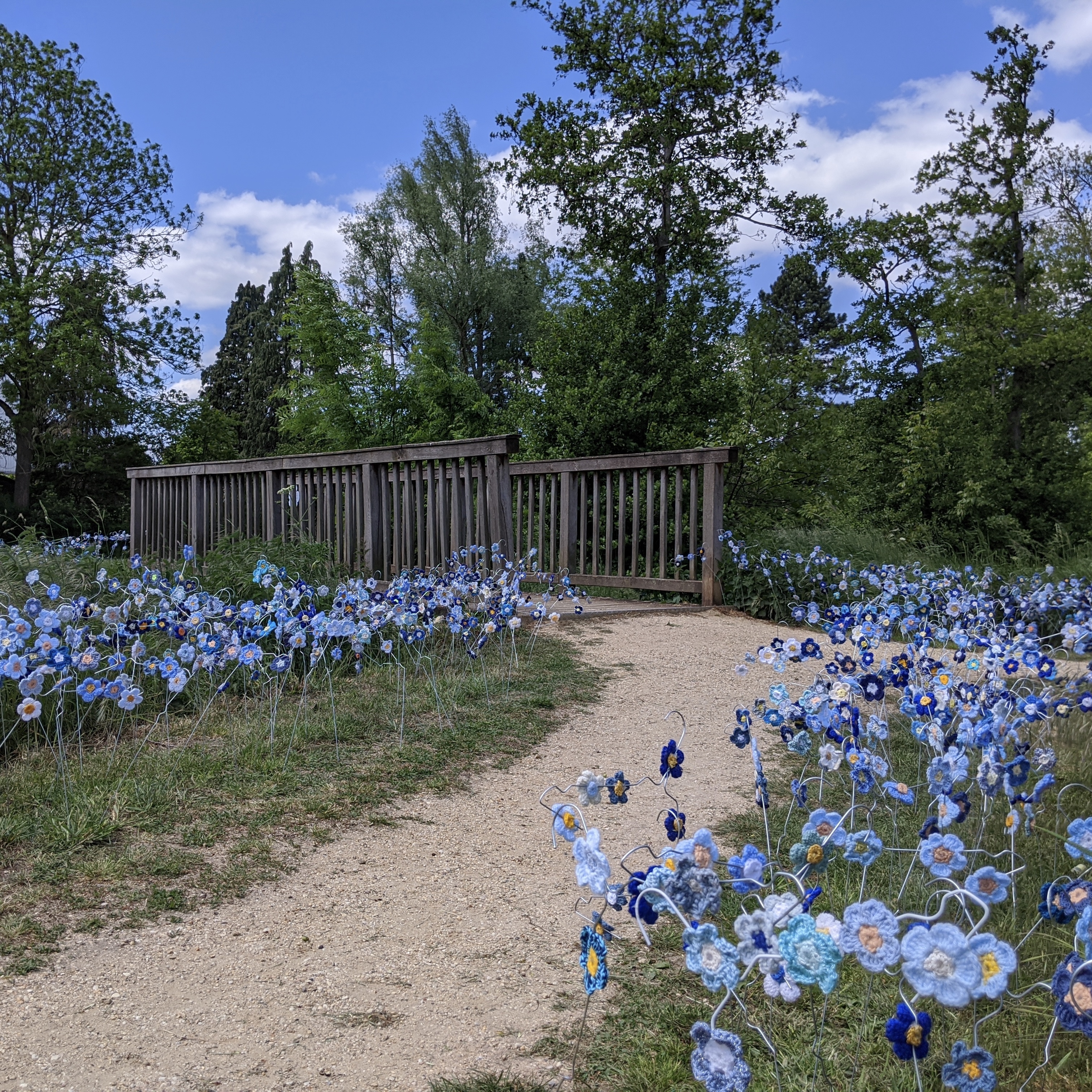 Ein Feld aus gehäkelten blauen Flachsblüten, im Hintergrund eine Brücke mit Holzgeländer